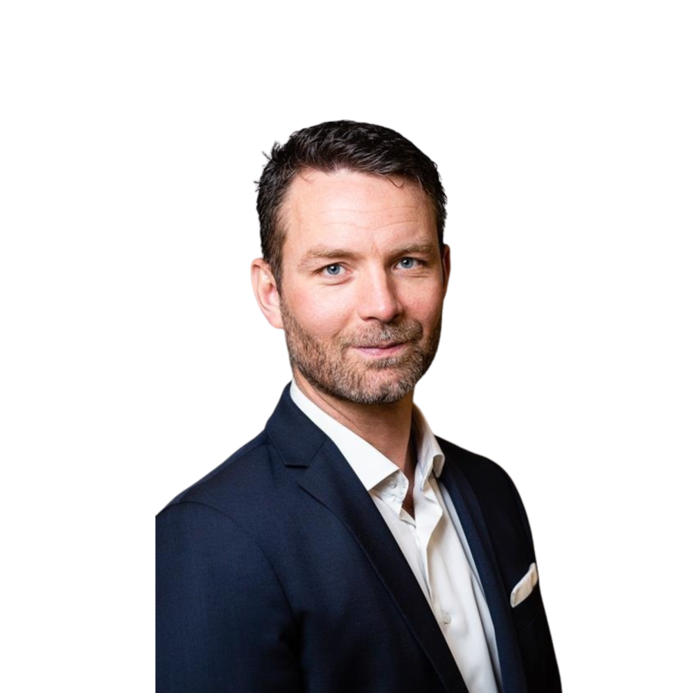 En mann med kort, mørkt hår og skjegg, kledd i en marineblå dressjakke, hvit skjorte og lommetørkle, står foran en hvit bakgrunn.