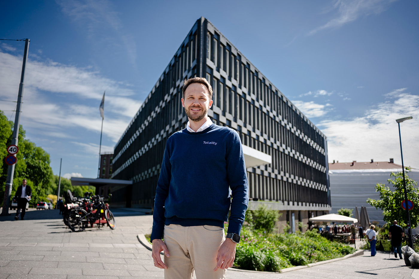 En mann i blå genser står smilende foran en moderne, kantete kontorbygning på en solrik dag.