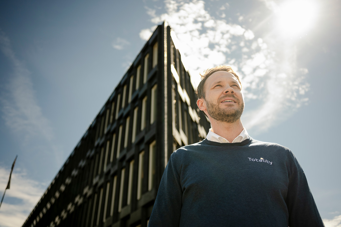En mann i en blå "Totality"-genser står utendørs foran en moderne bygning under en lys himmel der solen skinner.