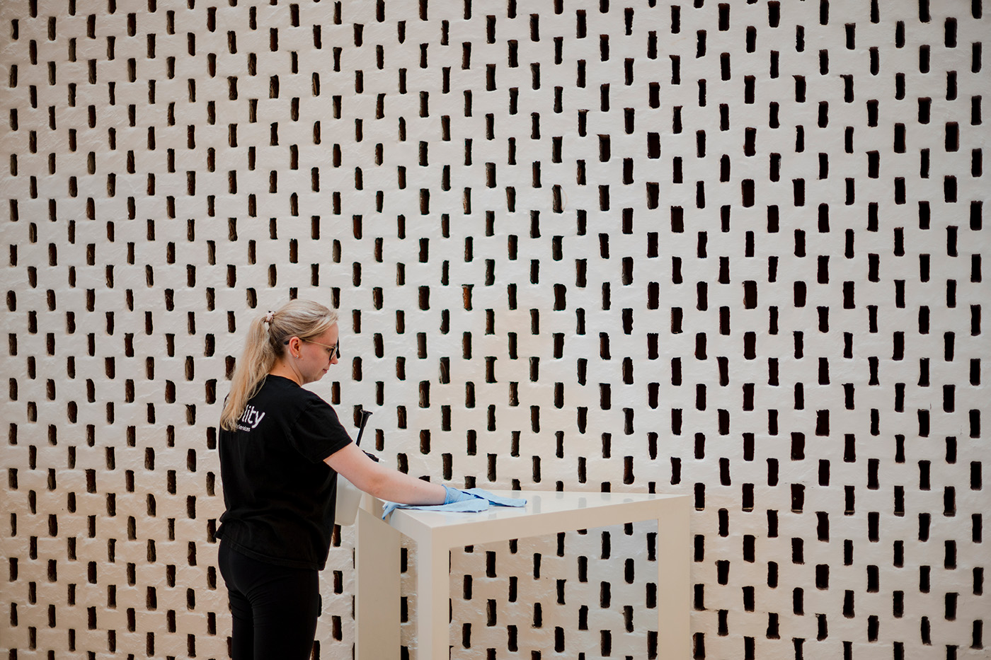 En person iført hansker og svart t-skjorte tørker støv av en stor, mørk skulptur i et godt opplyst innendørs rom.