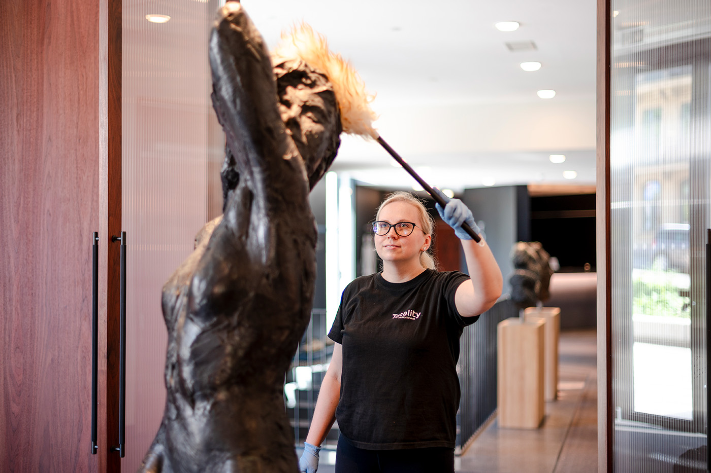 En person iført hansker og svart t-skjorte tørker støv av en stor, mørk skulptur i et godt opplyst innendørs rom.