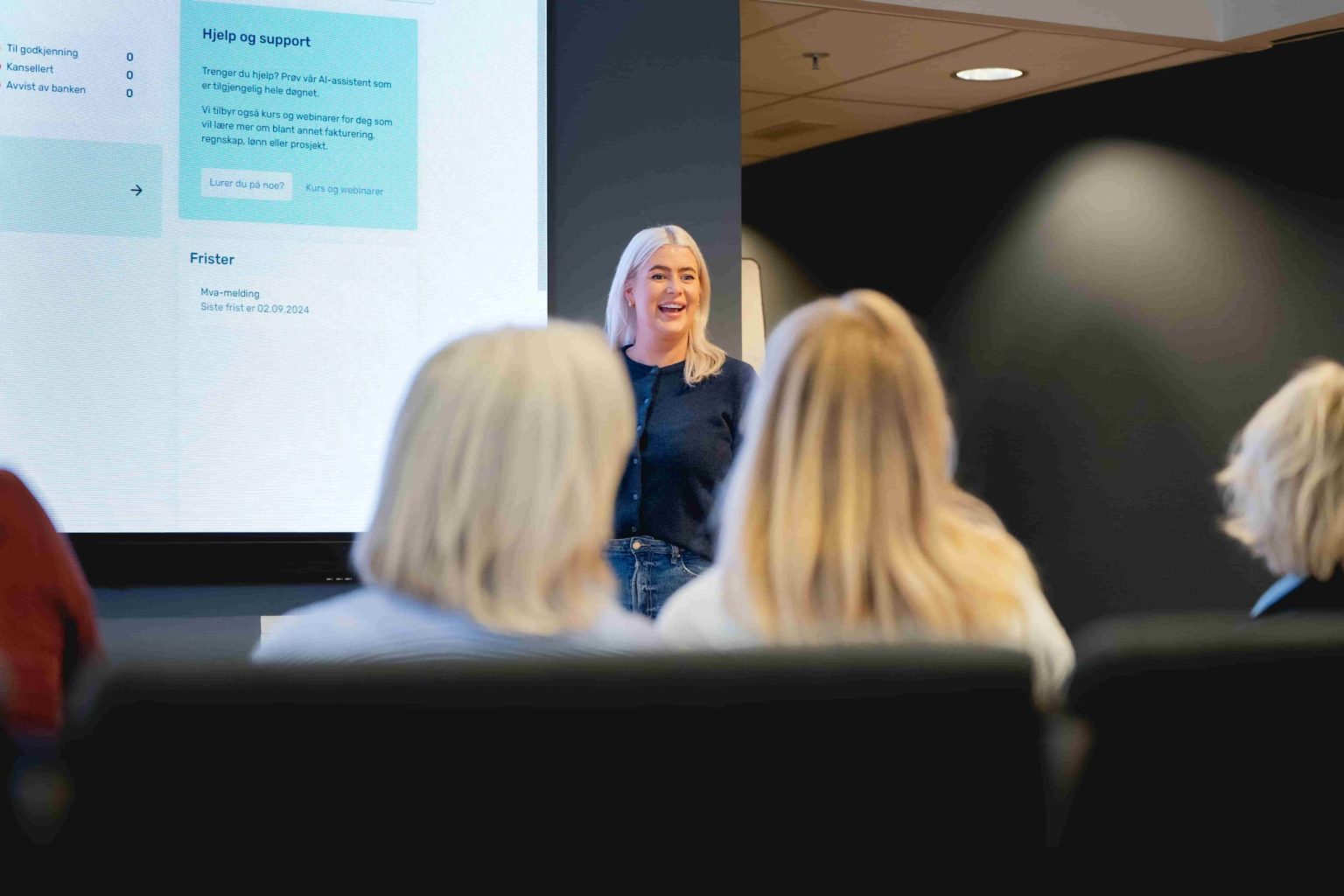 En kvinne står og smiler mens hun holder et foredrag for et publikum i et moderne konferanserom, med en stor skjerm som viser tekst bak henne.