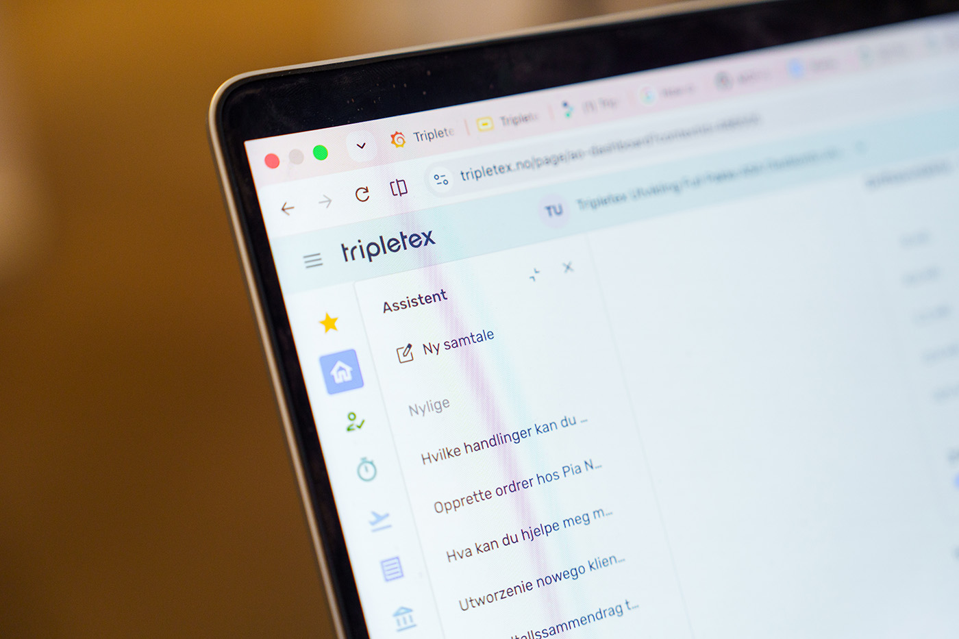 En mann i hvit skjorte står og smiler i et moderne kontor, med skrivebord, datamaskiner og kolleger synlige i bakgrunnen.