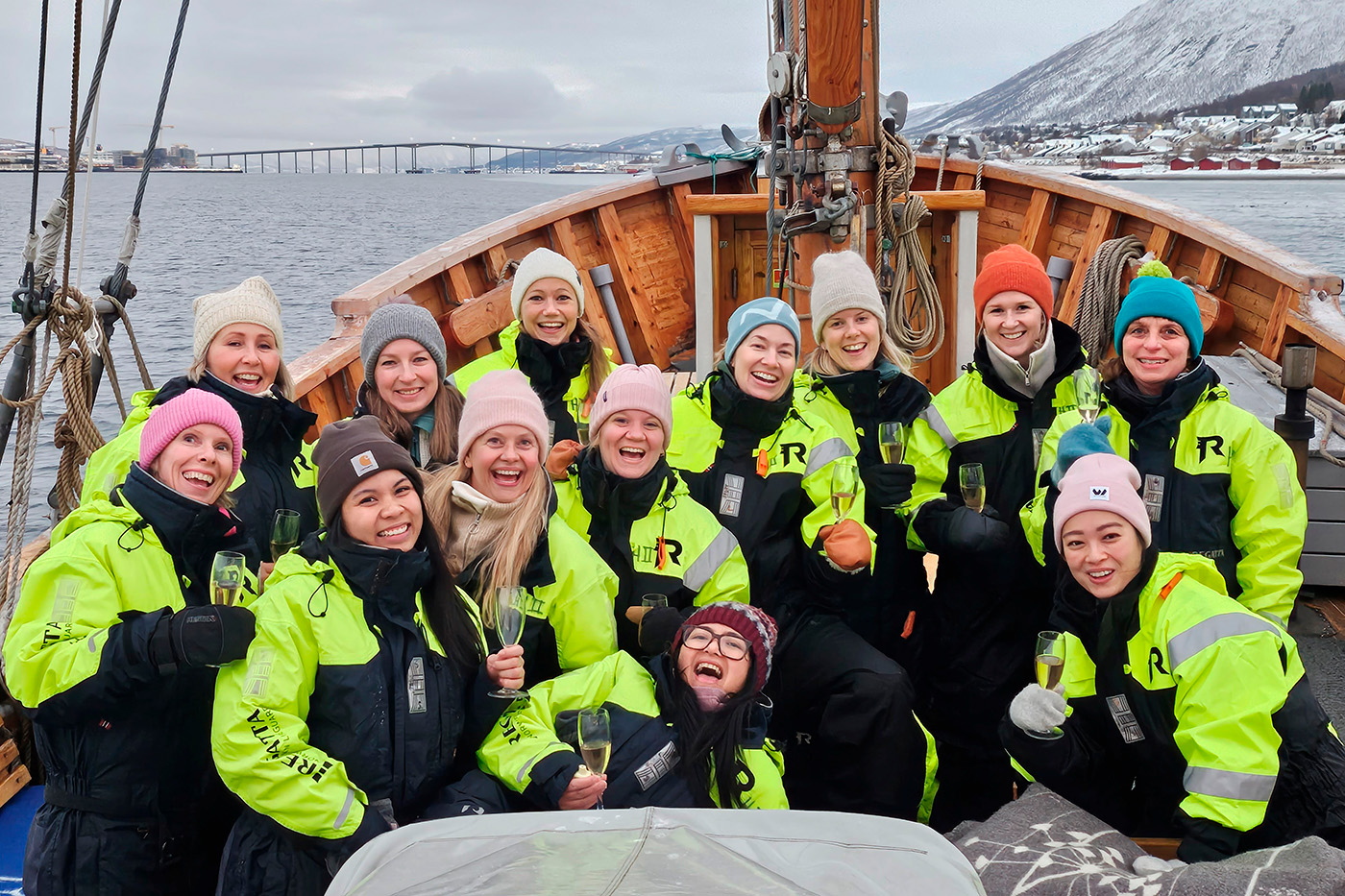 En gruppe på fjorten kvinner i knallgult og svart sjømannstøy nyter sine frynsegoder mens de skåler på dekket av en båt i en snødekt havn.