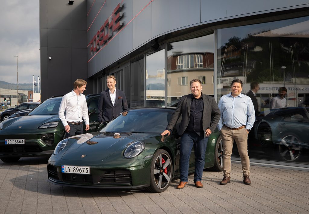Fire menn står ved to Porsche-biler som står parkert utenfor en bilforhandler.