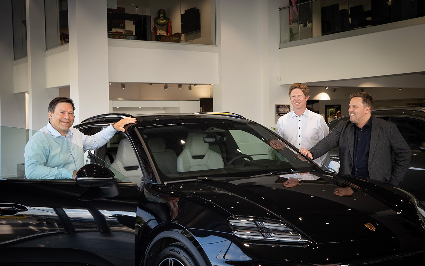Tre menn som står rundt en svart Porsche i utstillingslokalet til en bilforhandler.