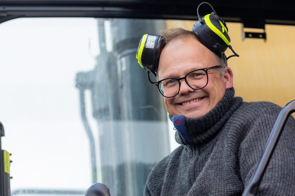 En smilende mann fra Ektvedt med briller og grå genser, iført hørselsvern inne i førerhuset på en maskin.