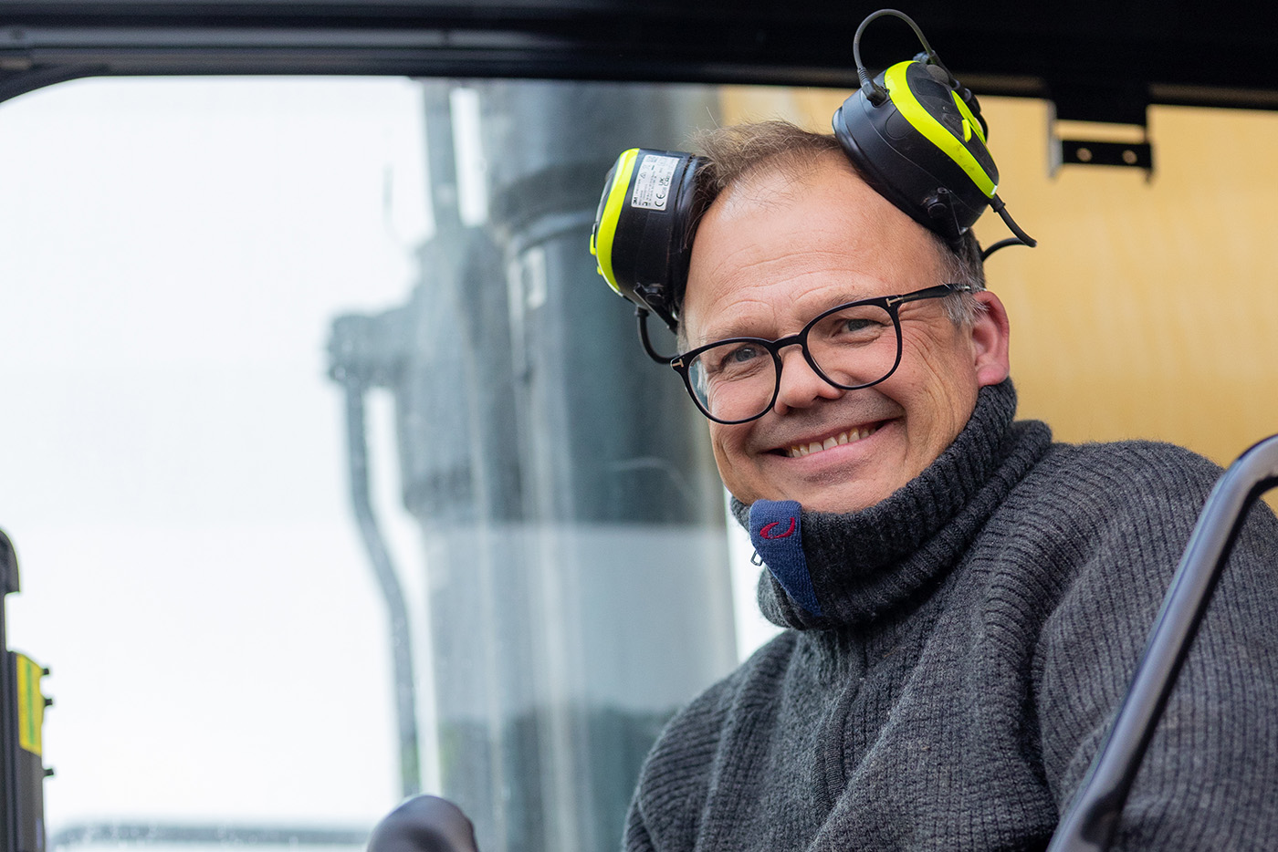 En smilende mann fra Ektvedt med briller og grå genser, iført hørselsvern inne i førerhuset på en maskin.